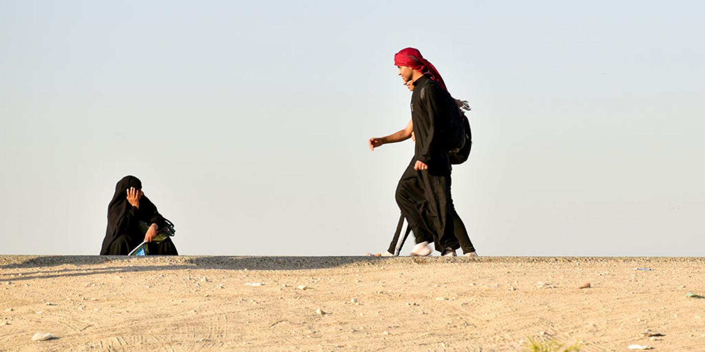 A black clad pilgrim squats and holds his head on the side of a desert road as other pilgrims walk along.