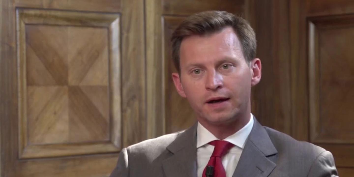 A man talks animatedly looking at the camera while sitting against a wood panelled wall.