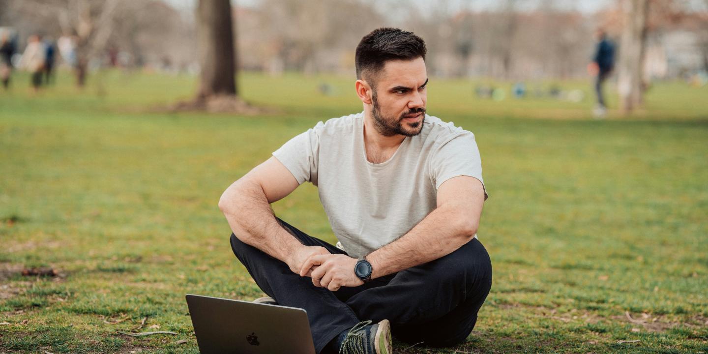 A man sits cross legged in a park with a laptop on the grass in front of him. He looks to one side.