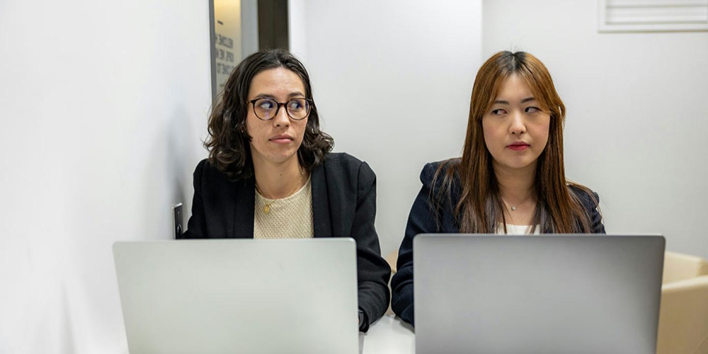 Two people sit beside each other working on laptops, One looks askance to the other who frowns.