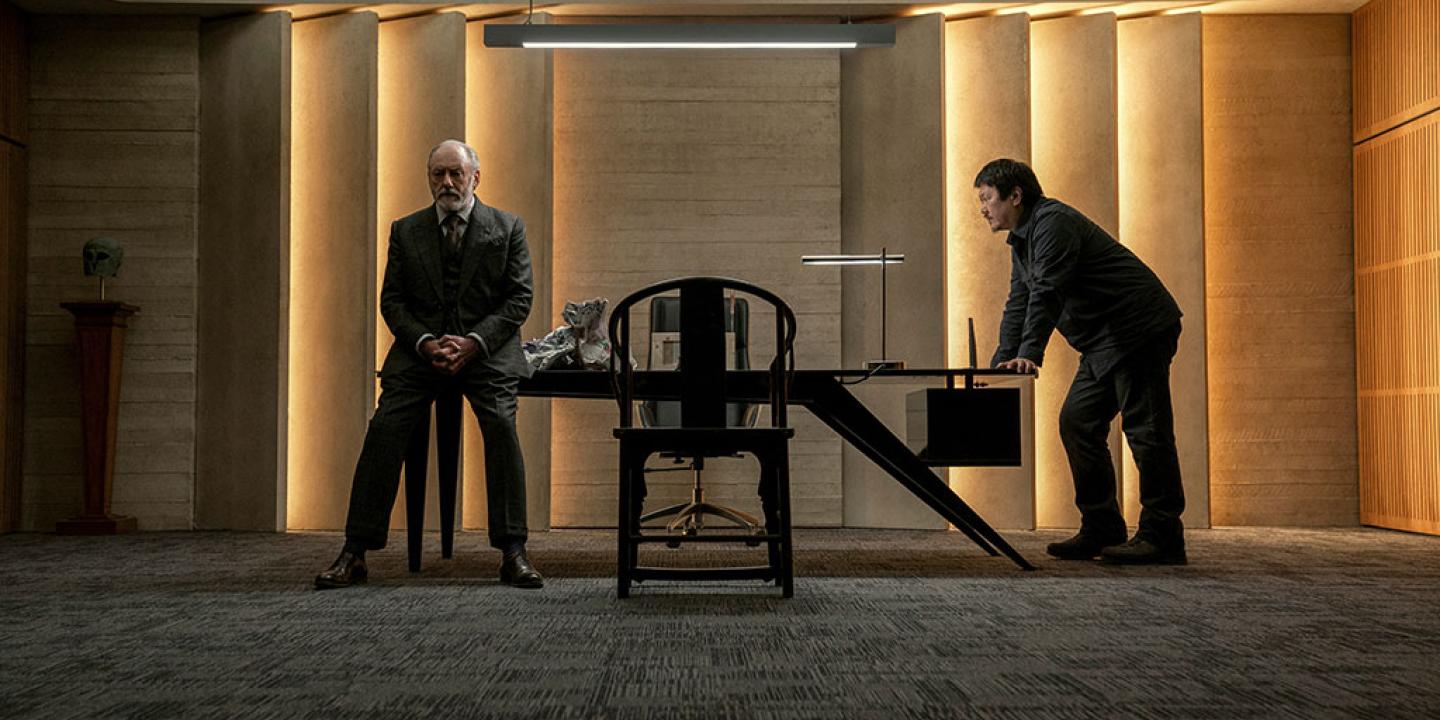 a man leans against one end of a table one another sits against its other end.