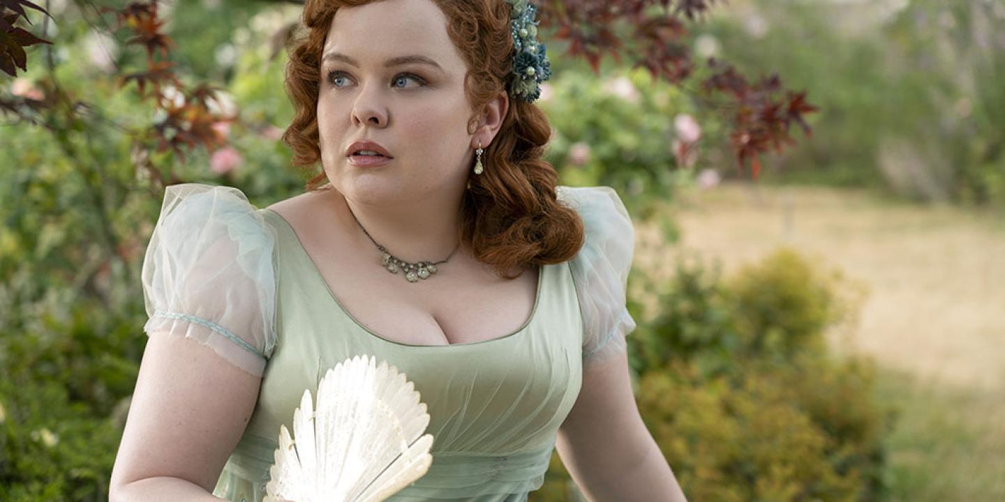 A young lady in Regency dress, holds a fan while looking around a garden.
