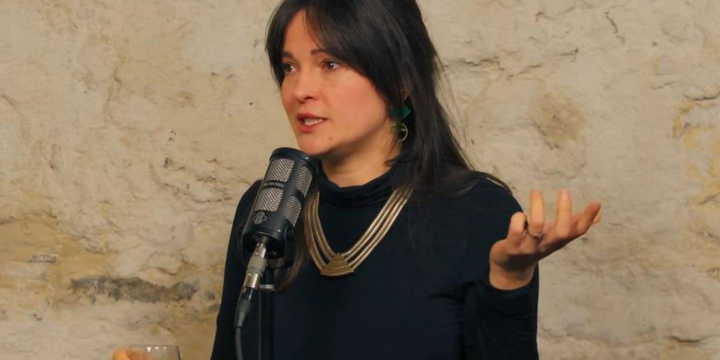 A woman sits a a table with a microphone in front of her, she is talking and gestures with one raise hand upward.