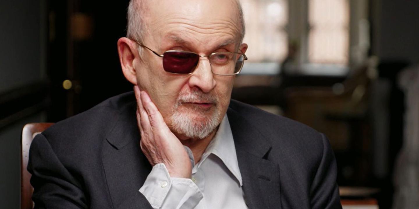 A man sits being interviewed and holds a hand to the side of his face, one lens of his glasses is tinted black.