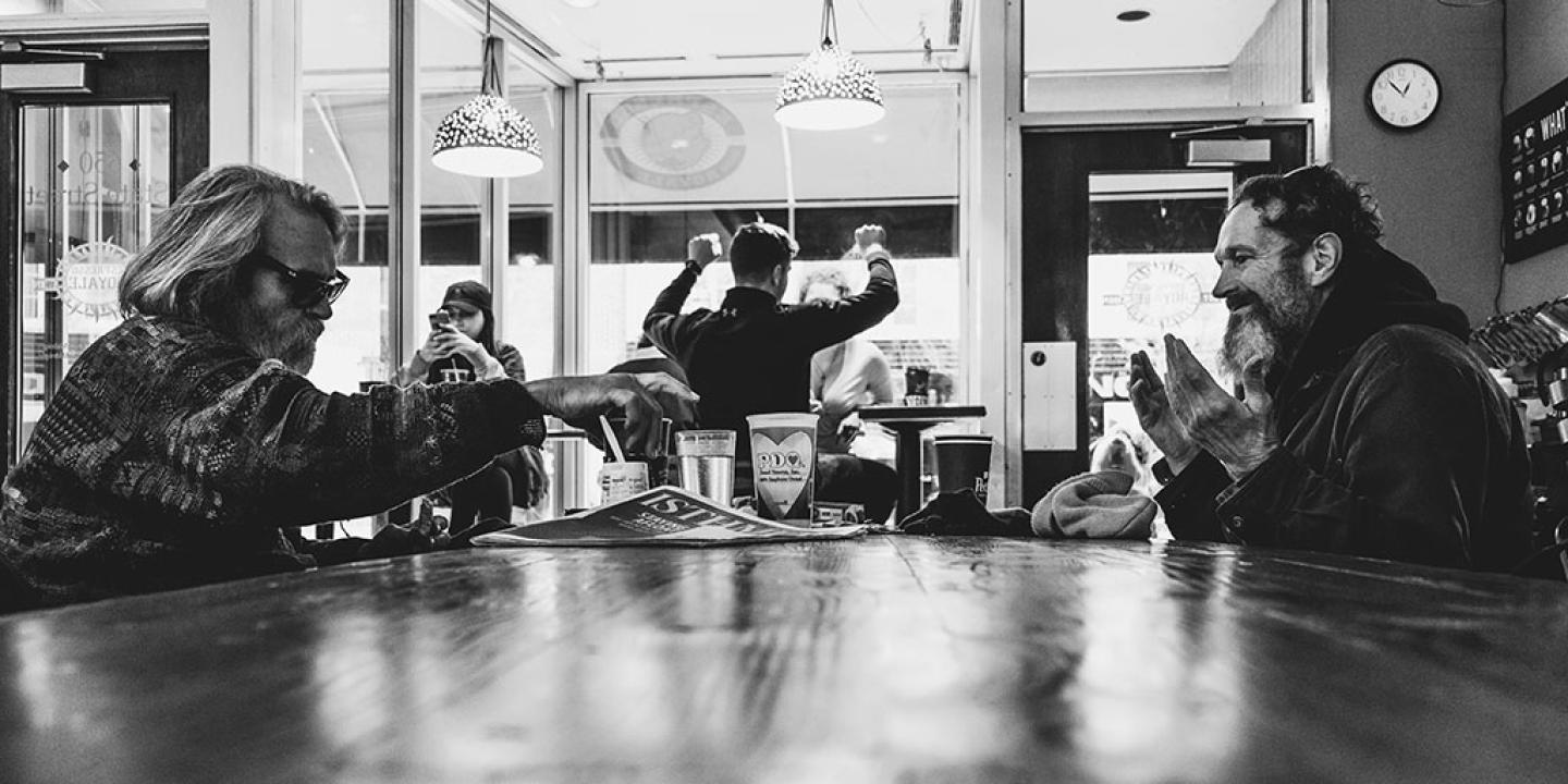 In a coffee shop, people sit around a table, one holds their hands up in surprise.