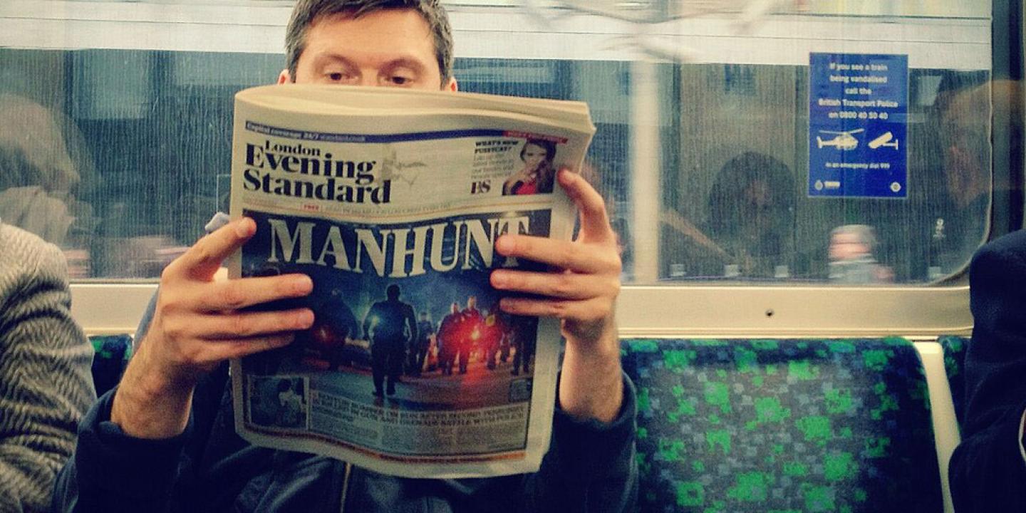 Seat on an underground train carraige, a passenger holds and reads a newspaper.