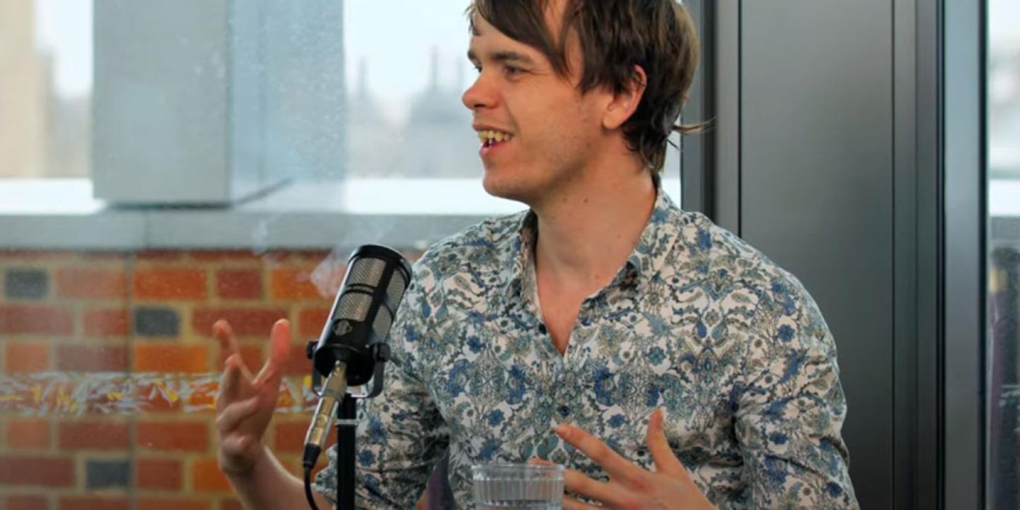 A podcast guest speaks in front of a mic and raises their hands to gesture.