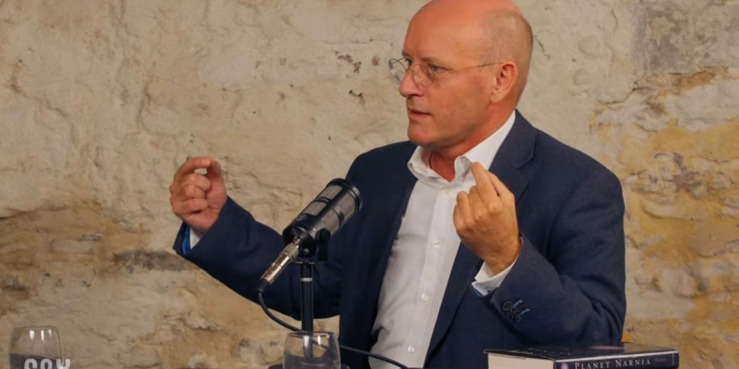 A man sits and speaks into a microphone and gestures with raised hands.