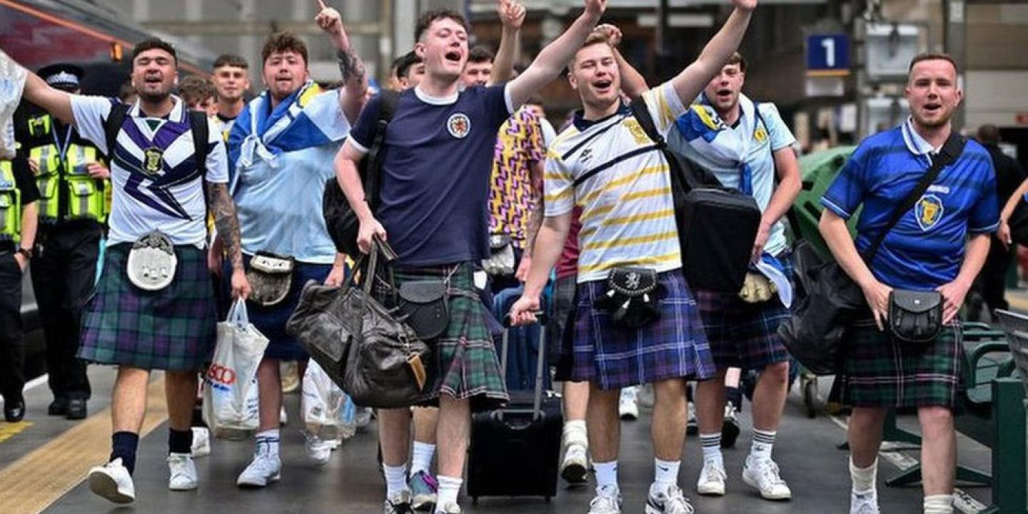 Scottish football fans wearing kilts march down a street singing and waving their arms alogt.