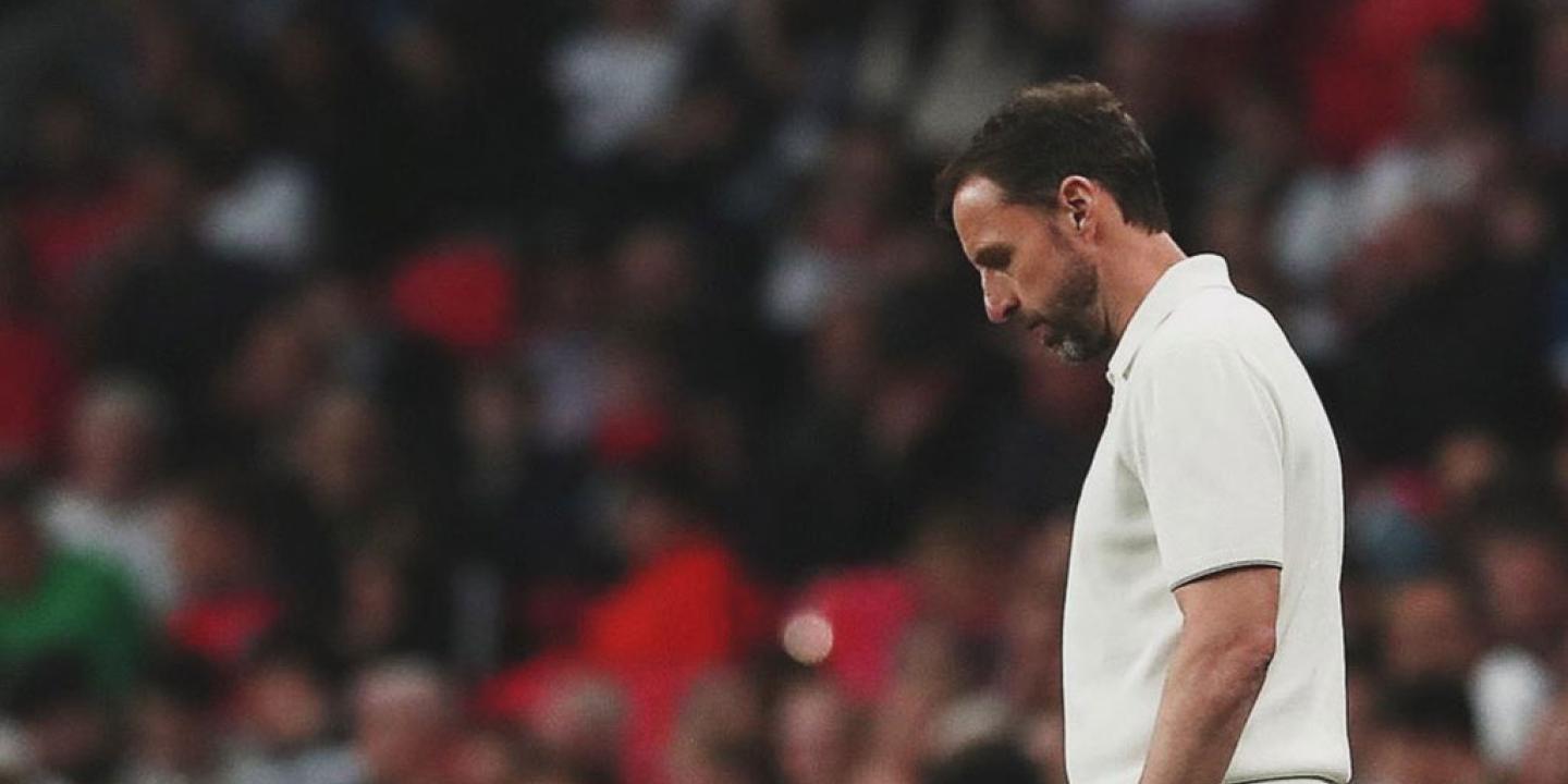 A dejected looking football manager ponders his feet while standing beside a pitch.