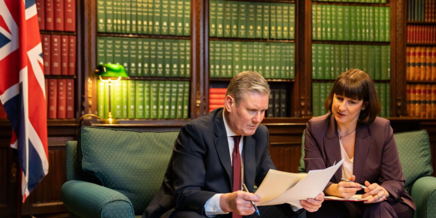 Kier Starmer sits on a sofa, leaning forward and holding papers he is reading. Rachel Reaves sits and looks on.