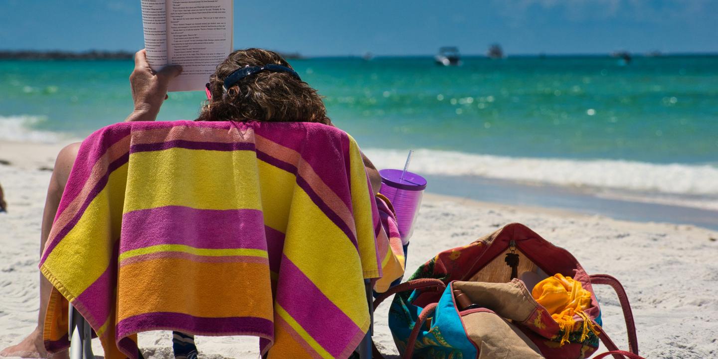 On a beach lounger someone holds a book aloft to read.