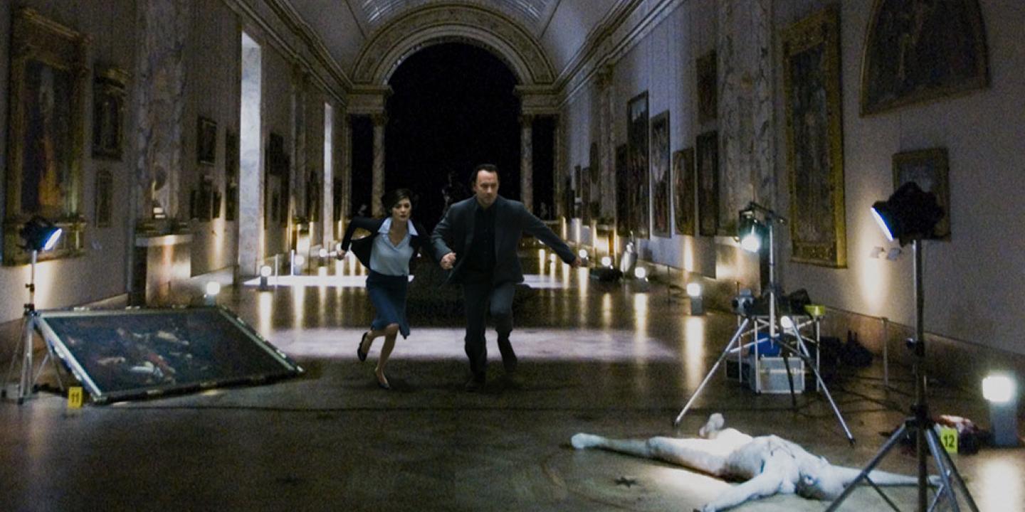 Two people run though a darkened art gallery towards a body lying amongst photography paraphanalia