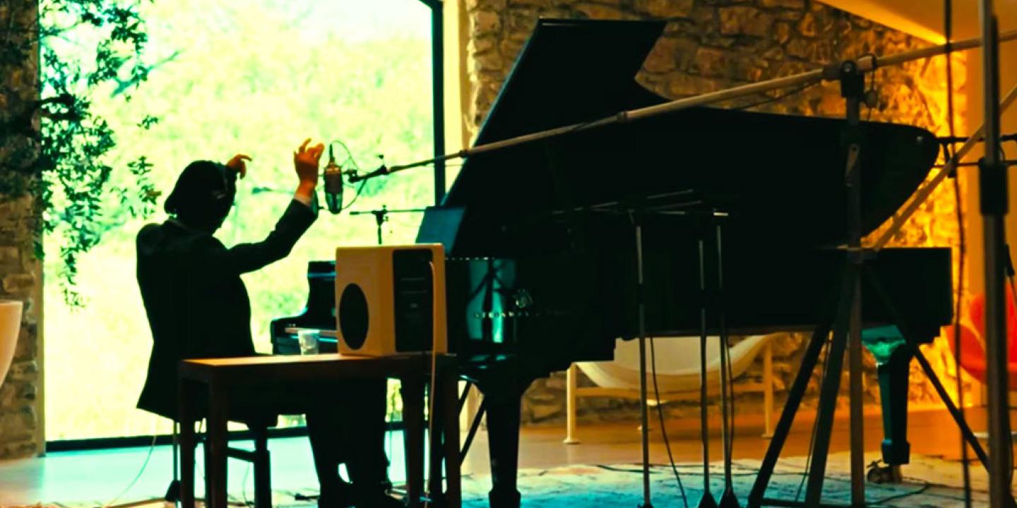A pianist raises his arms while sitting at a grand piano amid recording equipment.