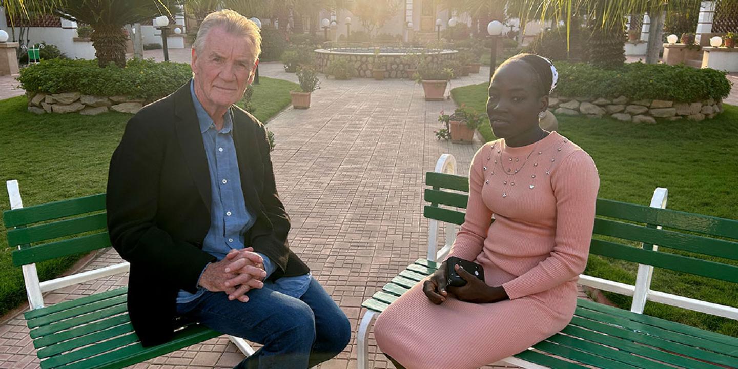 A TV interviewer sits across from a young woman in an outdoor setting