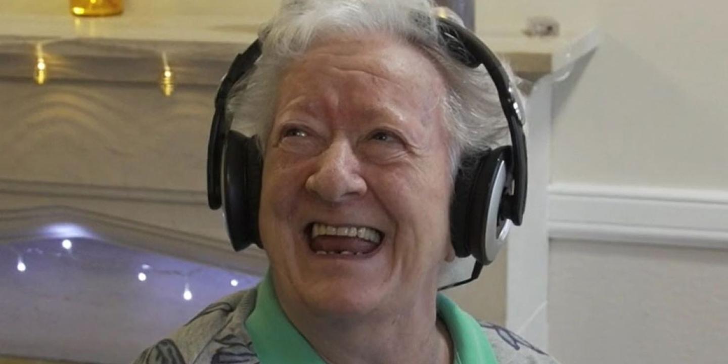 An elderly woman wearing headphone looks up and to the side with a big smile.