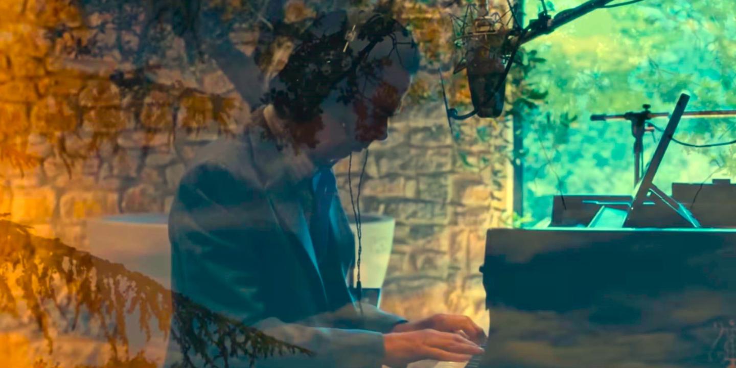 A pianist raises his arms while sitting at a grand piano amid recording equipment.