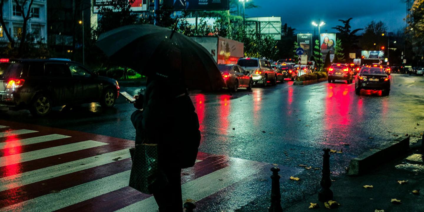 On a rainy night a pedestrian, holding a brolly, waits to cross a road.