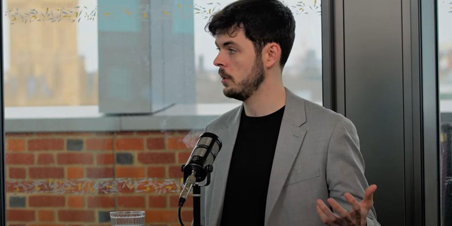 A podcast guest speaks in front of a mic and raises their hand to gesture.