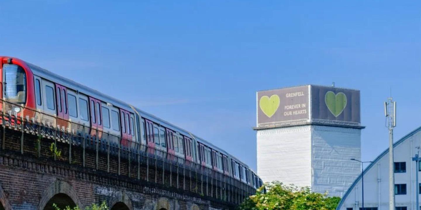 A tube train runs on a raised track, in the distance is a tower block wrapped in white material with a green heart on it.