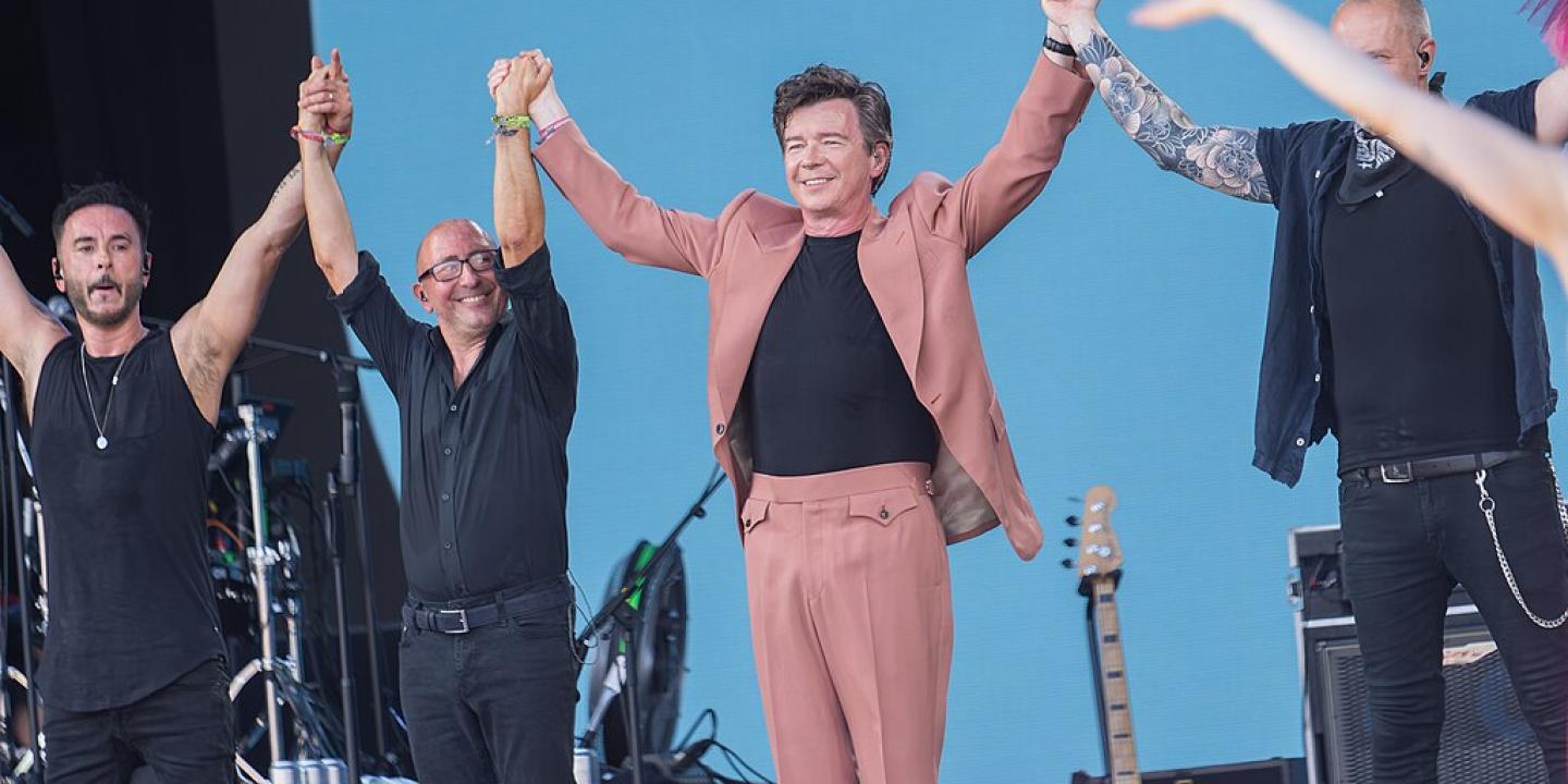 On a music festival stage, a popstar in a pink stages holds raised hands with his band.