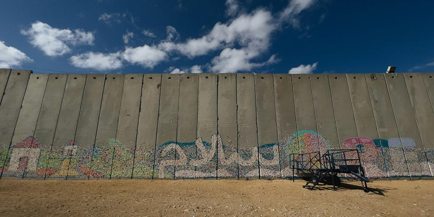 A graffitied concrete border wall stands below a blue sky and dusty ground