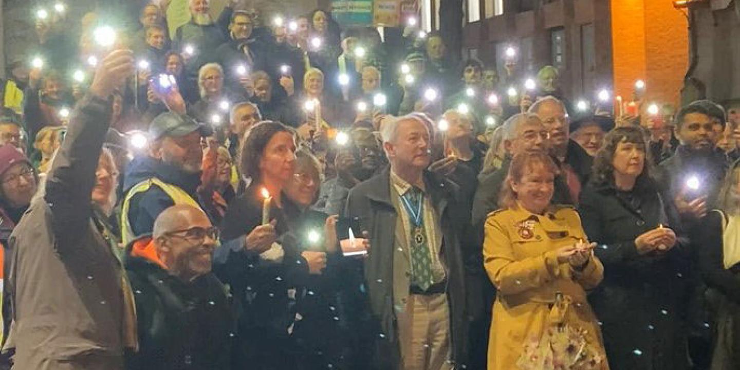 An outdoor vigil is lit by people holding up mobile phone lights.