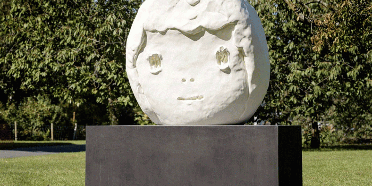 A large outdoor sculpture of a round child-like head sits on a dark plinth.