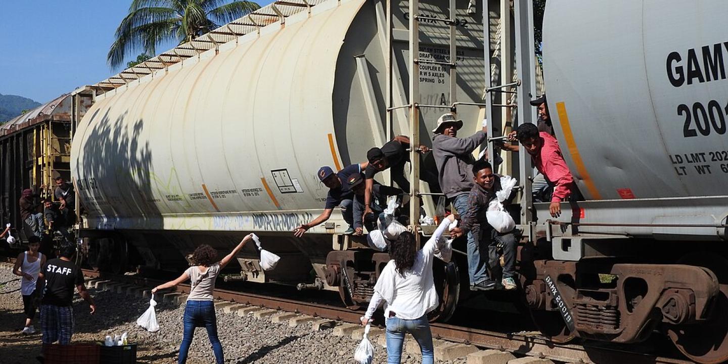 Migrants on a freight train reach for food bags held aloft by people on the track side.