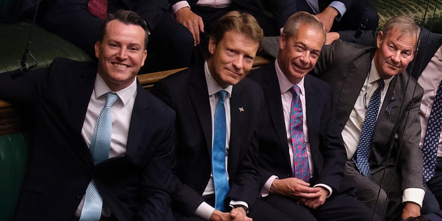 Four men in suits, sit next to each other smiling, in the House of Commons.