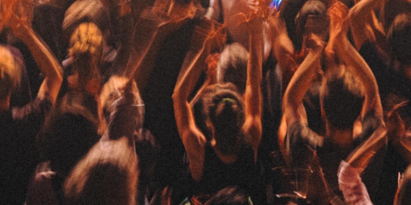 Concert goes stand, bathed in warm light, raising their hands in the air, facing away from the camera to the stage