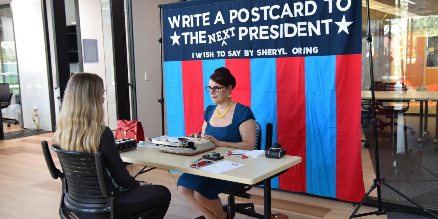 Two people sit at the table, one dictates as the other types. Behind a banner reads: write a postcard to the next president.i