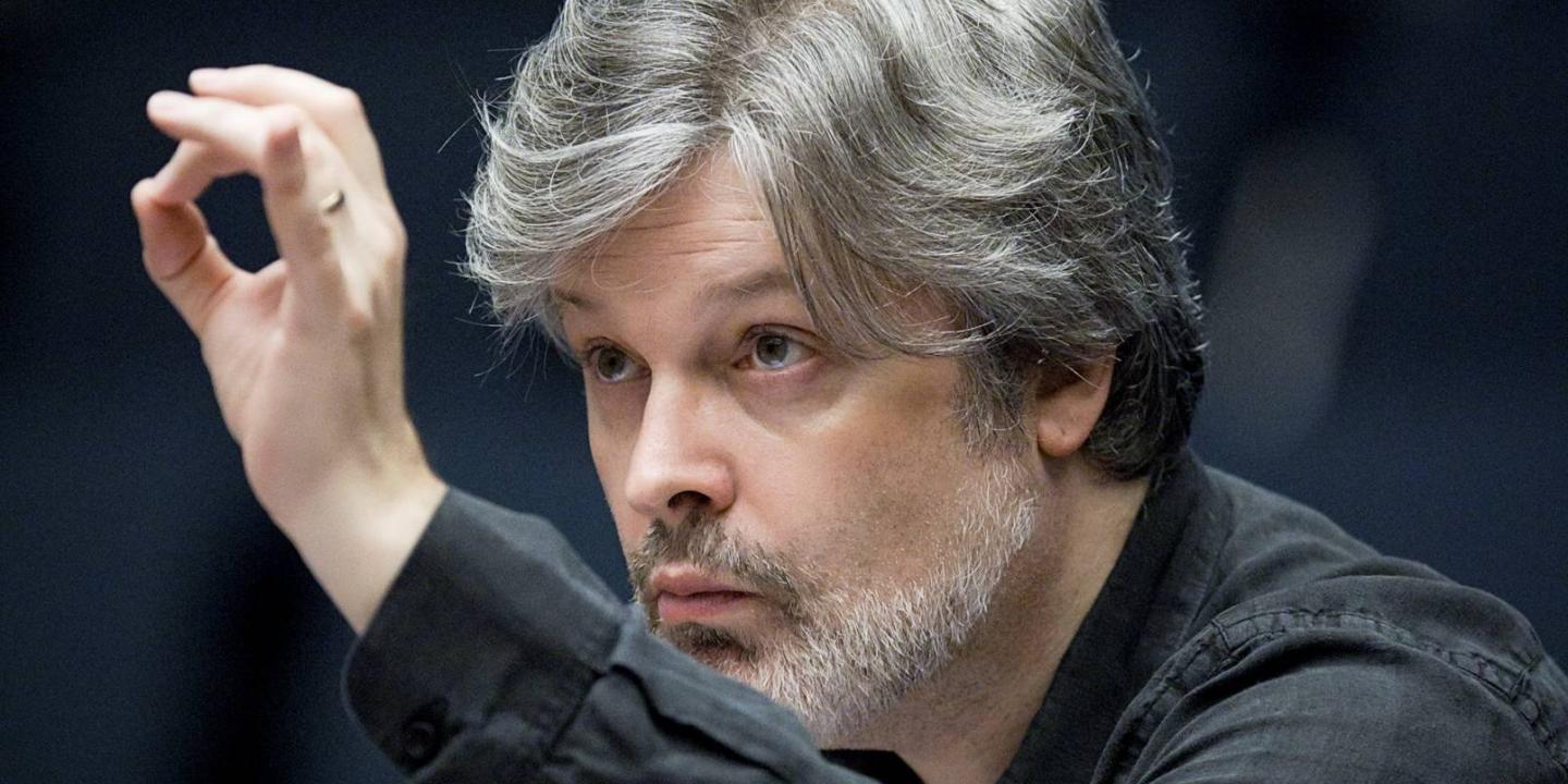 A conductor leans in toward an unseen orchestra with a raised hand.