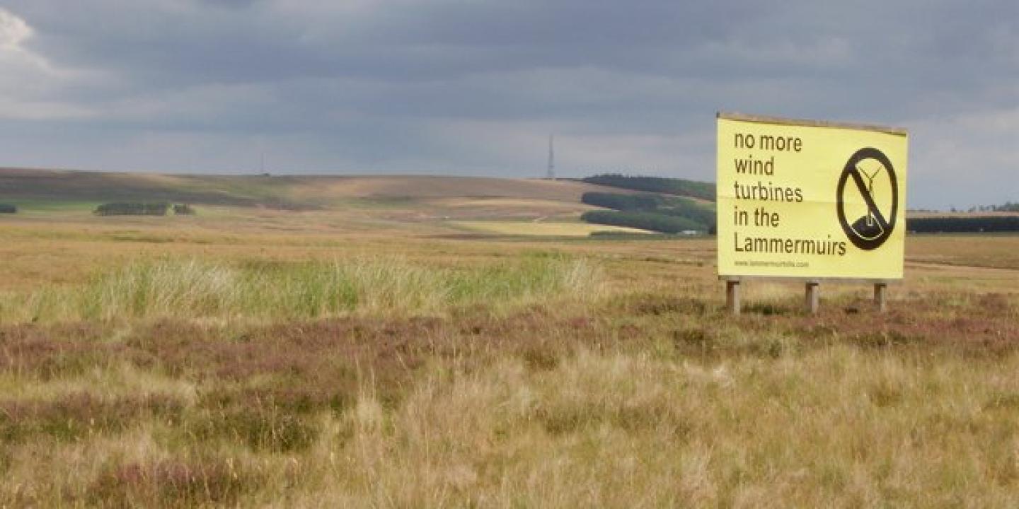 A sign protesting a proposed wind farm stands on flat moorland.