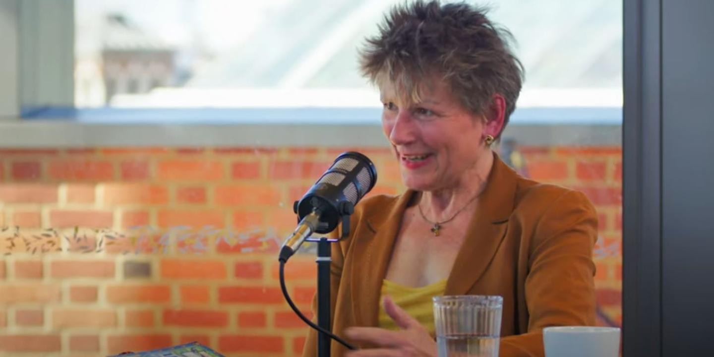 A woman sitting at a table in front of a microphone talks and gestures