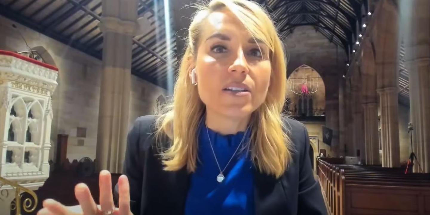 A woman sitting in an empty church talks to the camera and gestures with one hand.