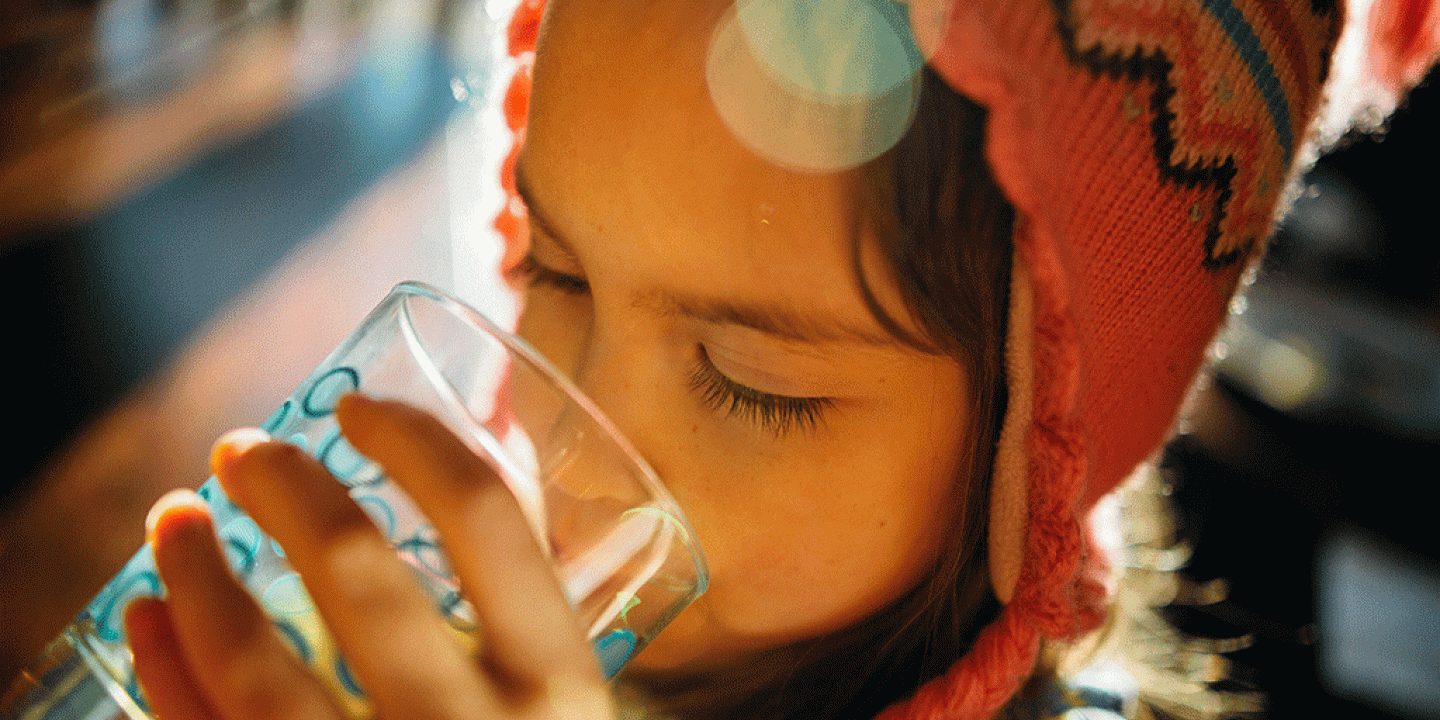 A child wearing a wool hat holds a glass and drink water from it.