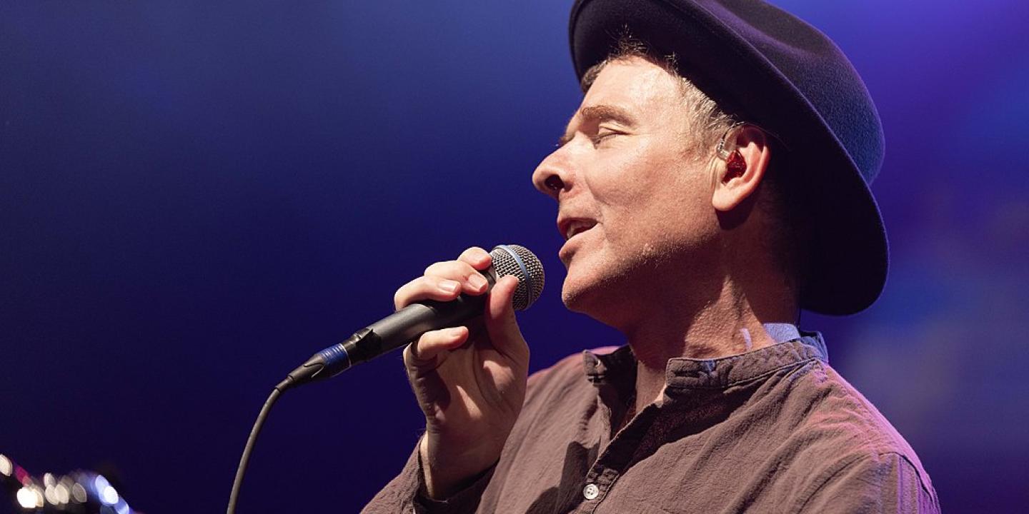 A singer, wearing a hat, pulls his head back holding a note, and a mic.