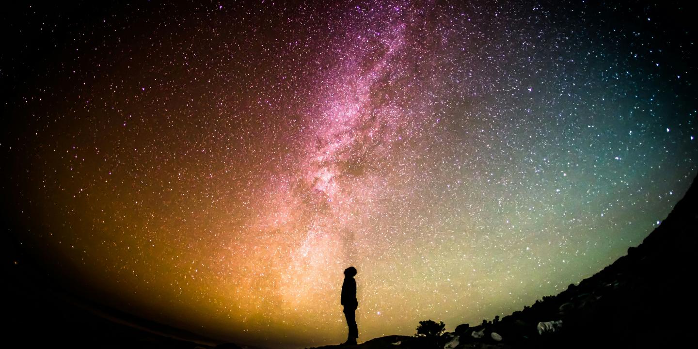 A fish eye lens view of a person standing silhouetted, looking up to a colourful night sky with the Milky Way across it