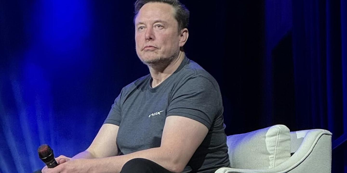 Elon Musk, wearing a t-shirt slouches forward, holding a mic, while sitting on a stage chair.