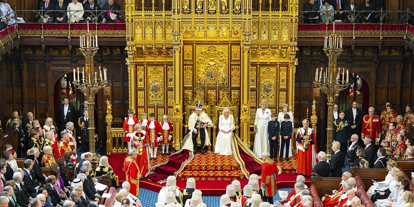 A wide angle picture shows a king and queen on thrones before many people in ceremonial clothes.