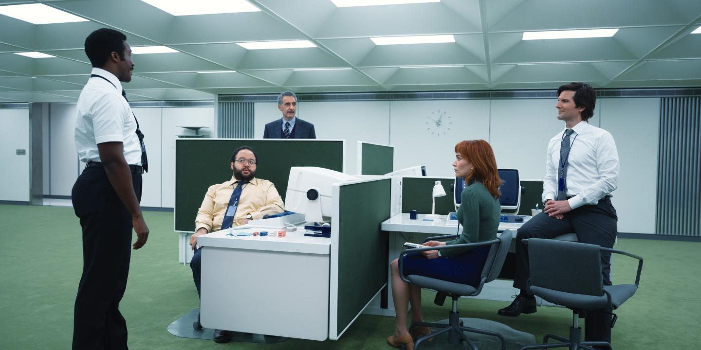 In a retro-future styled office, workers stand or sit around a pod of desks.