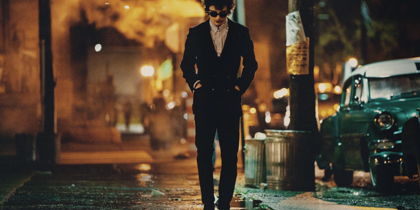 A hunched man wearing dark glasses and a dark suit, walks down a New York Street at night, with his hands in his pockets.