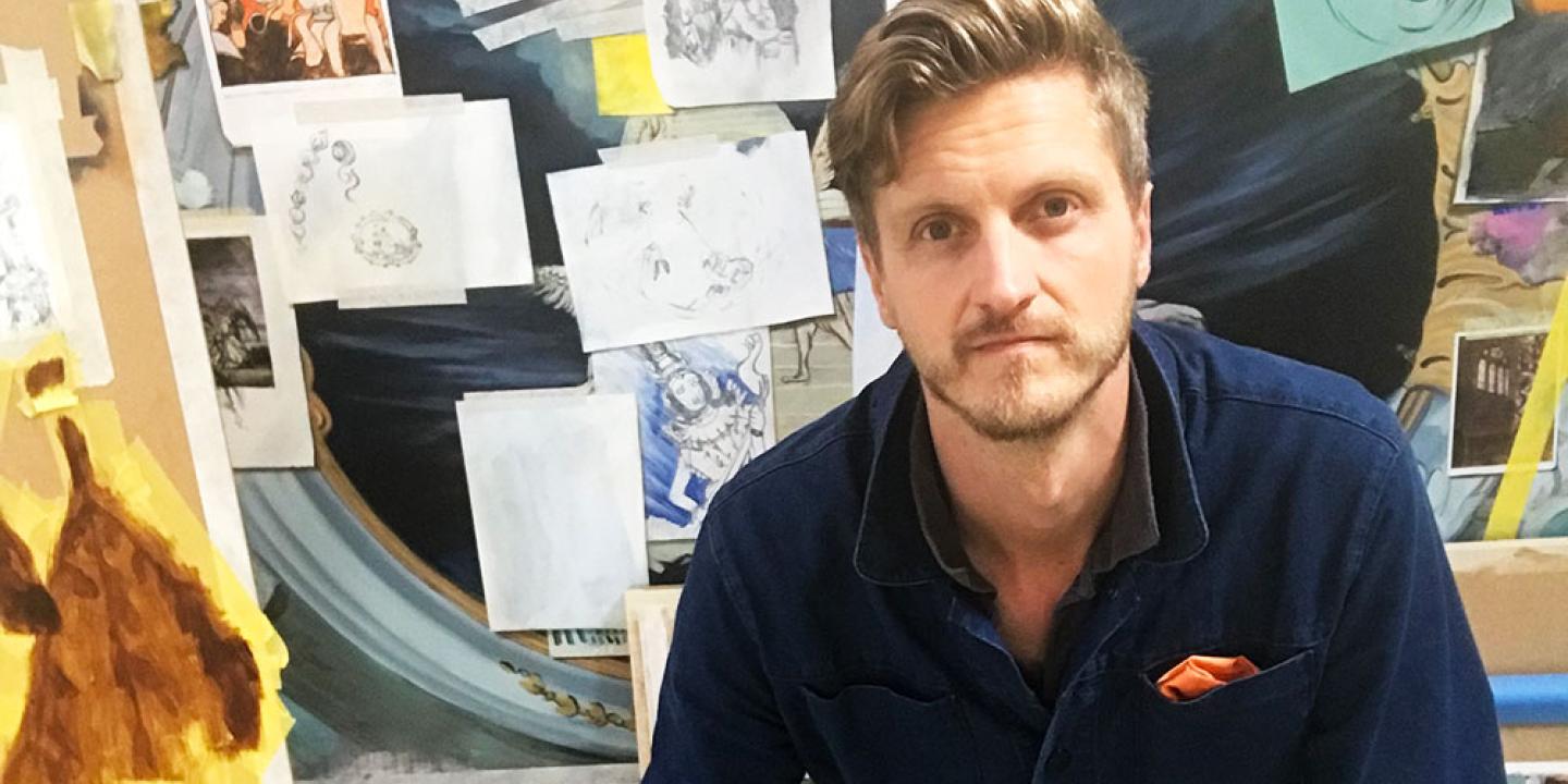 An artist sits in front of a board covered in images, canvases and paper.