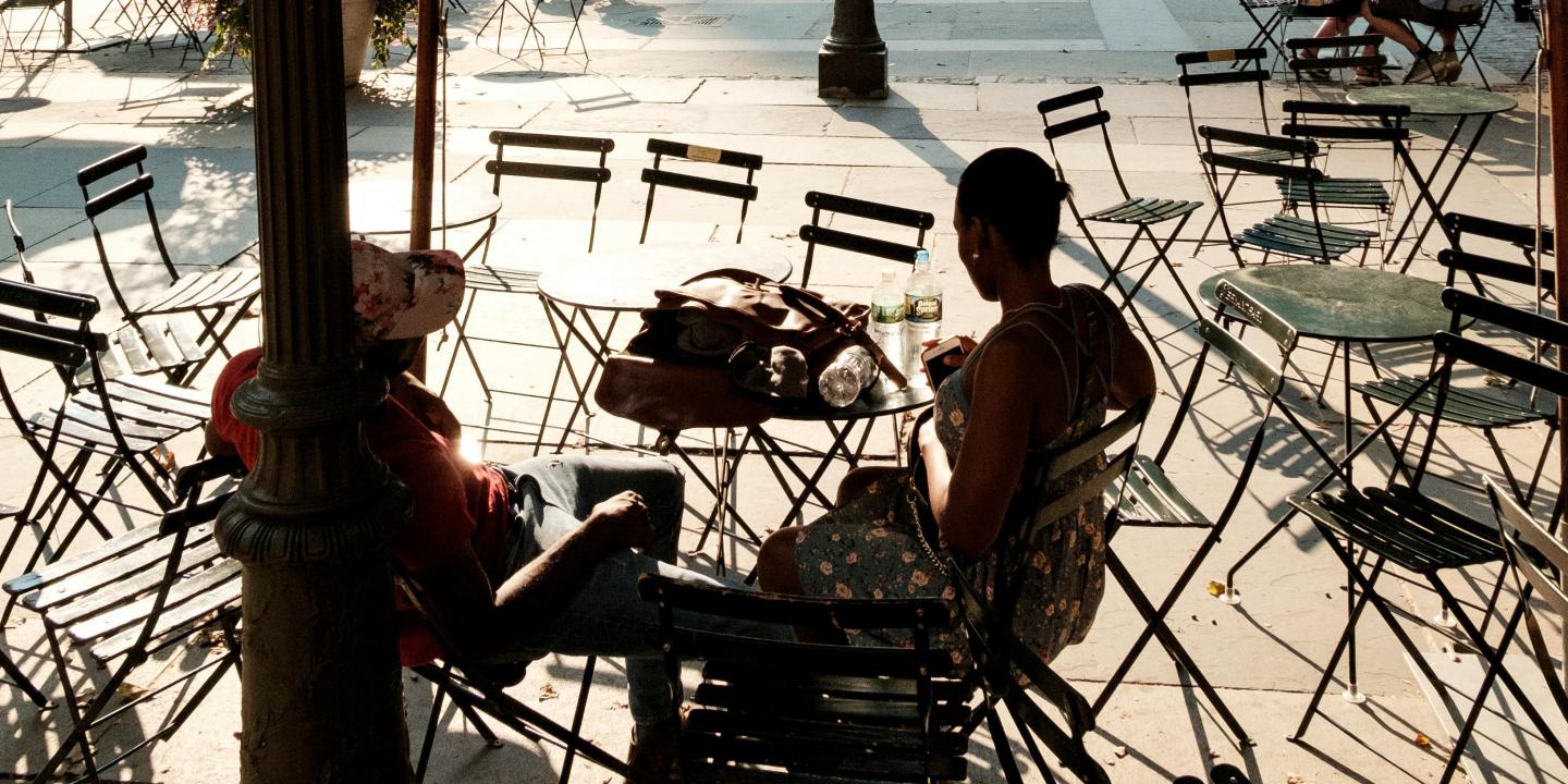 Two people. sitting at a street cafe amid empty tables and chairs, are silhoutted.