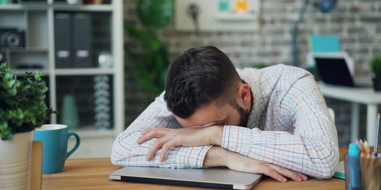 A man lies asleep on a closed lap top on a desk.