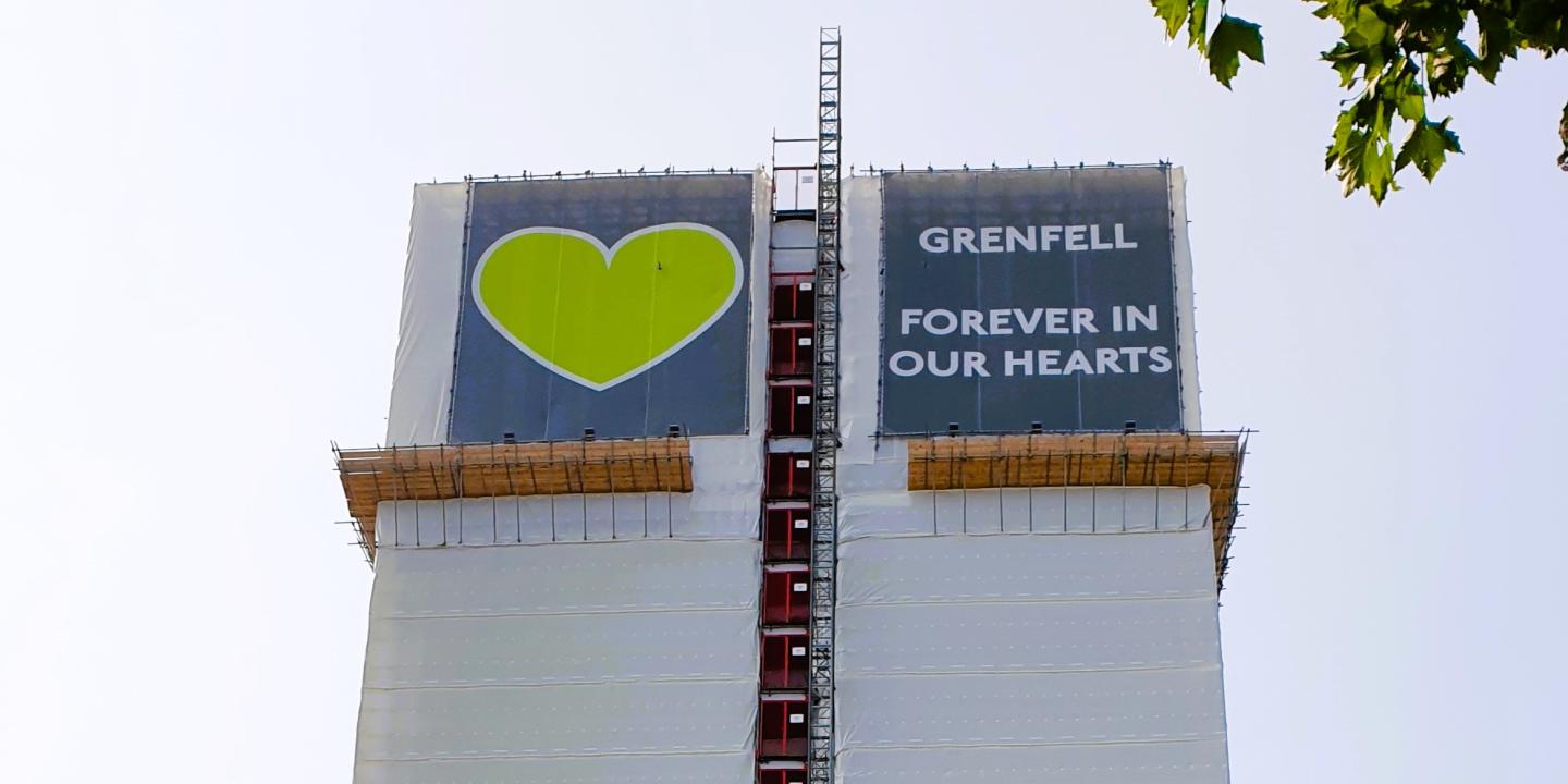 A white building wrap around a tower is topped by a green heart and the slogan: Grenfell Always In Our Heaets.