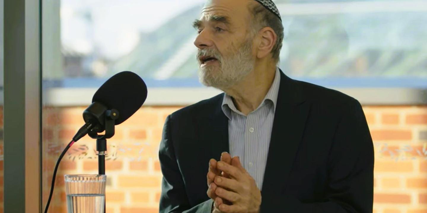 A podcast guest holds his hands together in front of himself as he raise his head in speech.