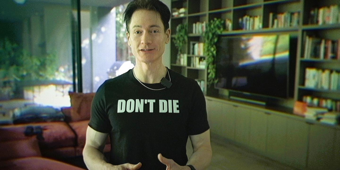 A man stands in his home wearing a black t-shirt that reads 'DON'T DIE'.