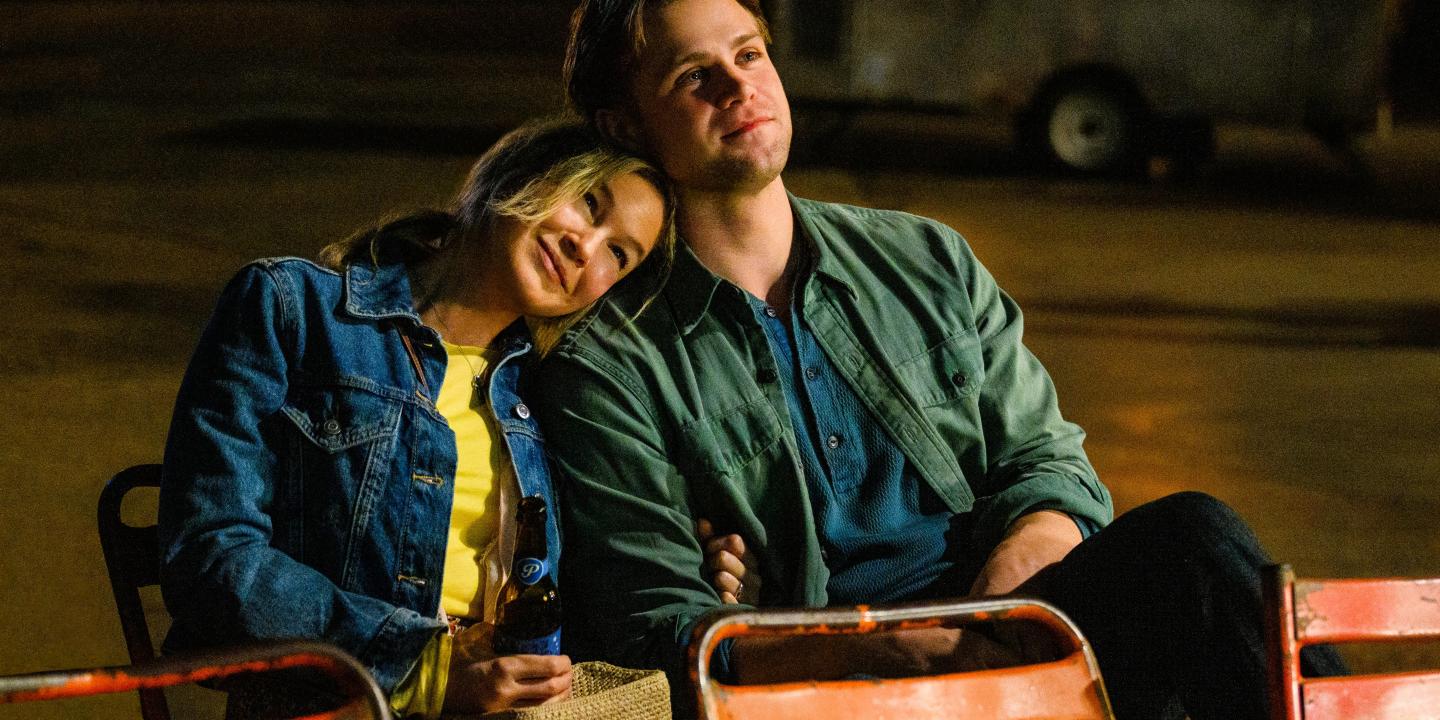A couple sit on outdoor seats, her resting her head on his shoulder.